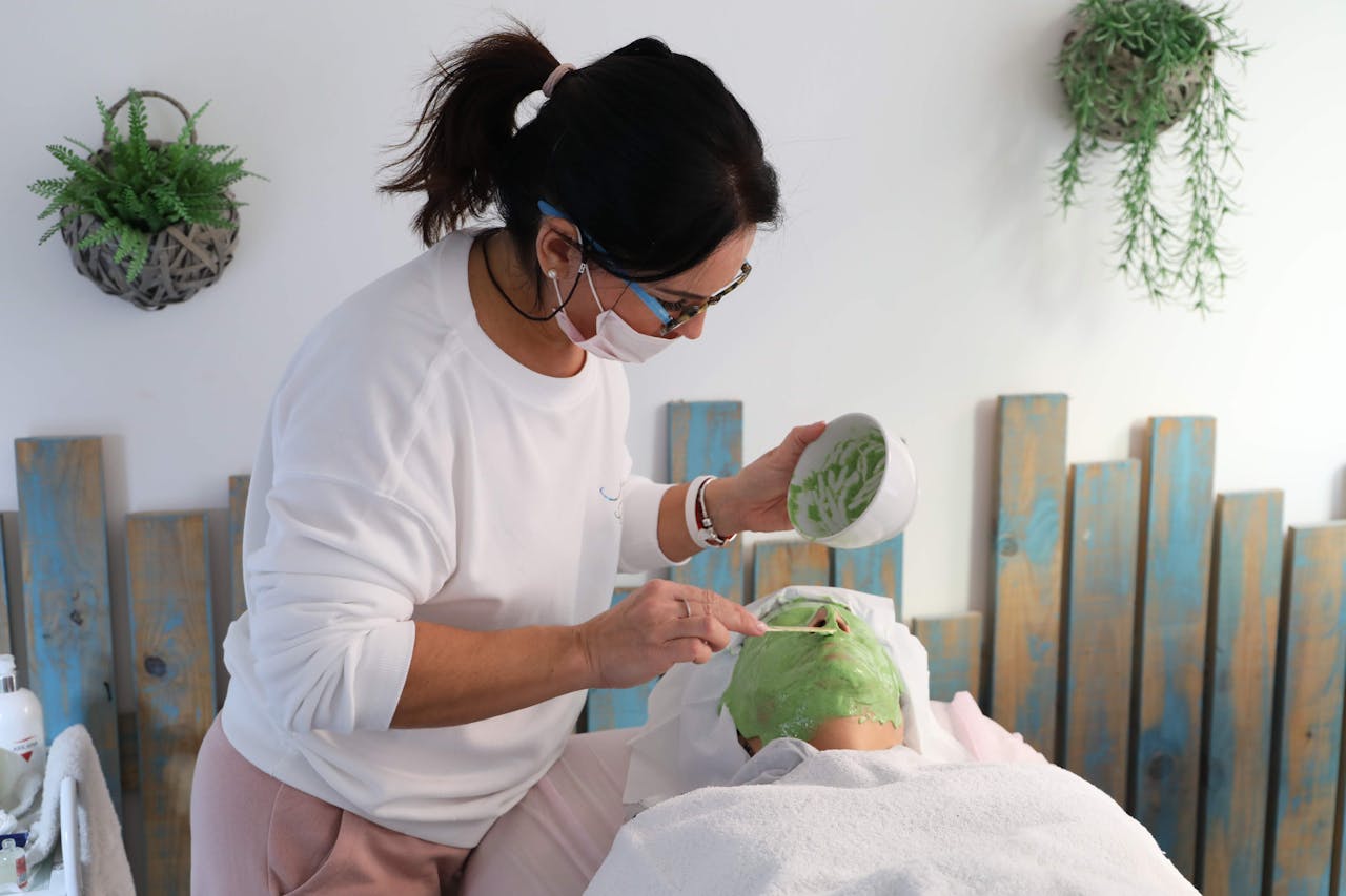 Aesthetician applying a green clay mask to a client during a relaxing spa facial treatment.